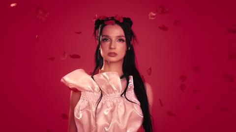 Elegant woman with black hair and flower headpiece poses in a pink gown against a red studio backdrop with floating rose petals.