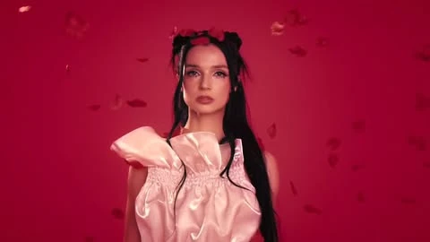 Elegant woman with black hair and flower headpiece poses in a pink gown against a red studio backdrop with floating rose petals.