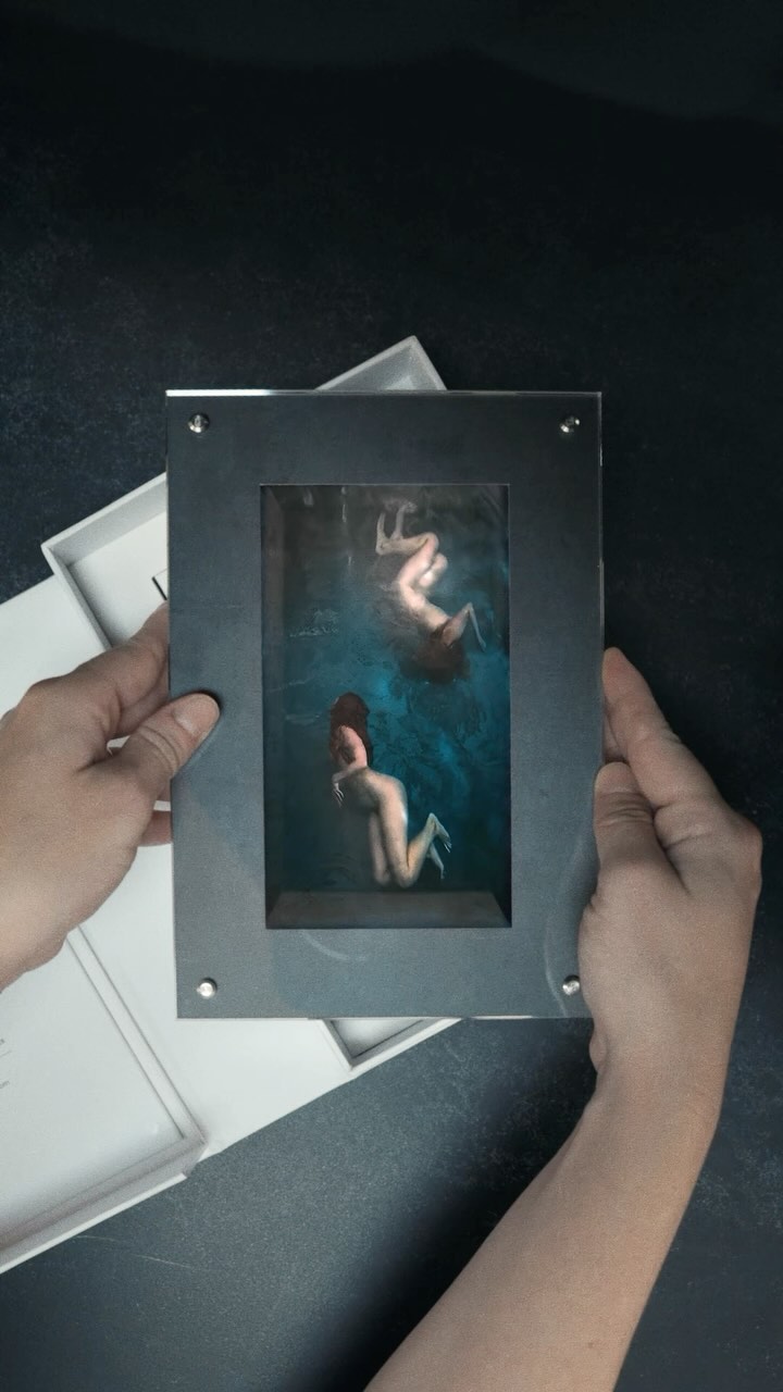A silver framed photo of two nude women in a pool is held up with a white box and dark background. Soft lighting with a slightly muted color palette.