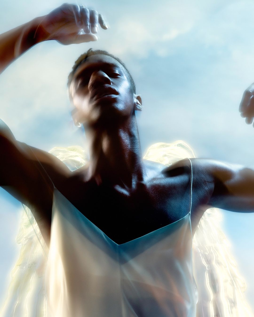 Striking studio shot of a muscular African American male with angel wings, wearing a white tank top against a blue sky with soft, blurred focus.