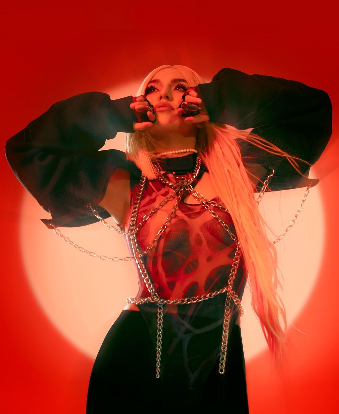 Blonde woman in red-toned studio setting. Woman has fair skin, and is posing with a graphic red and black top, and black bottoms.