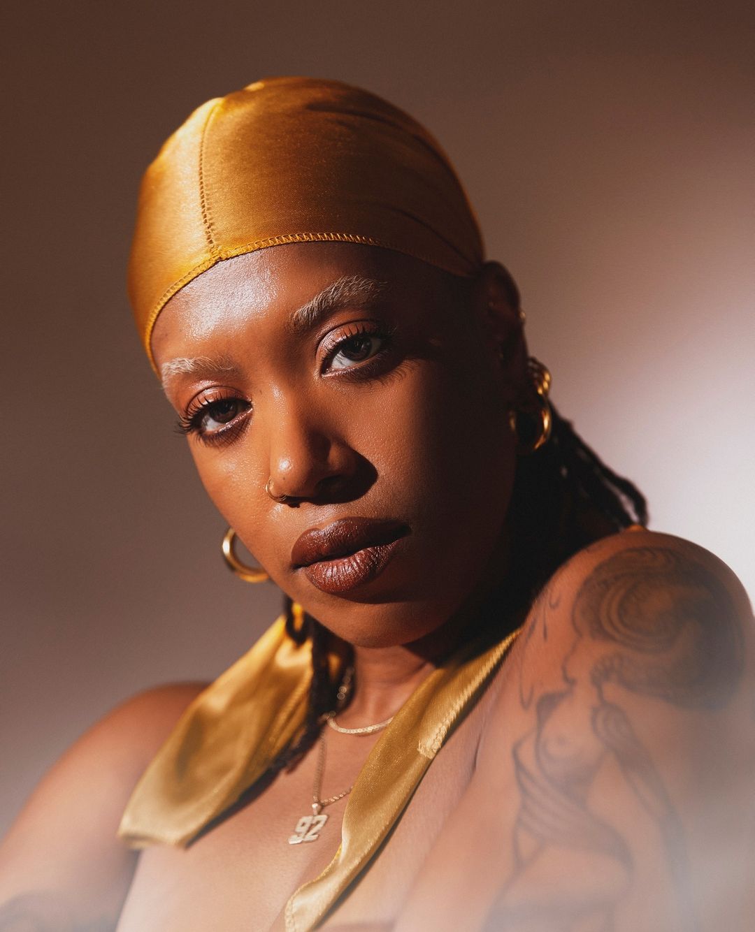 Close-up portrait of a Black woman with gold headscarf looking directly at the camera with neutral expression in warm studio lighting.