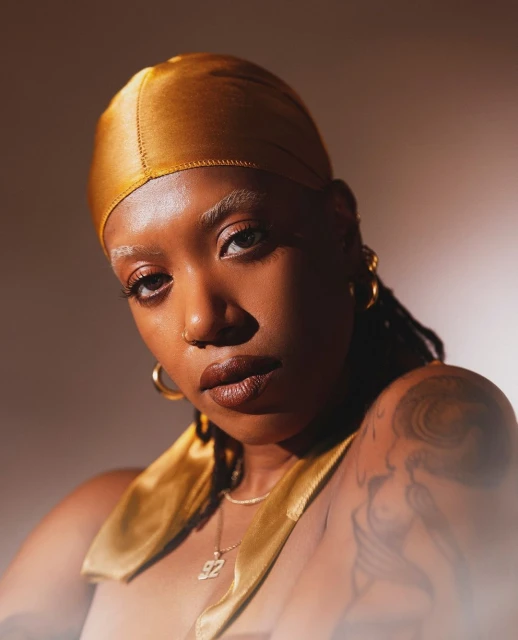 Close-up portrait of a Black woman with gold headscarf looking directly at the camera with neutral expression in warm studio lighting.