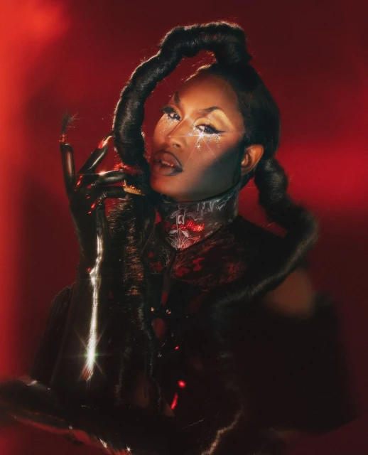 Woman with elaborate makeup and hairstyle poses in a studio with red lighting. She wears black gloves and a textured garment.