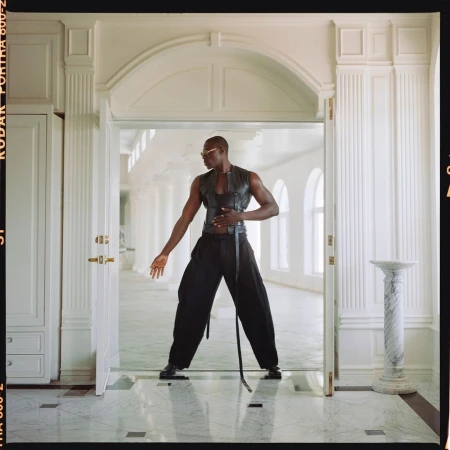 Fashion shot of a muscular man in a black leather vest and baggy pants standing in a white doorway of a bright white hallway.