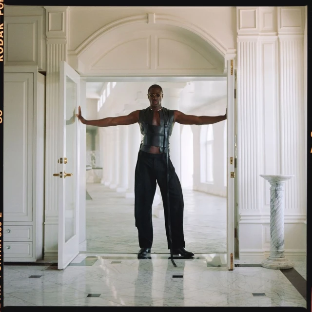 An adult male stands confidently in a doorway, wearing a leather vest and trousers, inside a bright, white, elegant hallway with marble floors.