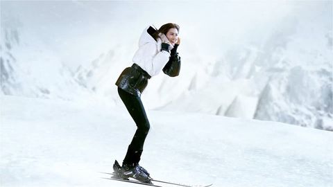 A woman in a white puffer jacket and black pants is skiing down a snow-covered slope against a backdrop of snow-capped mountains.