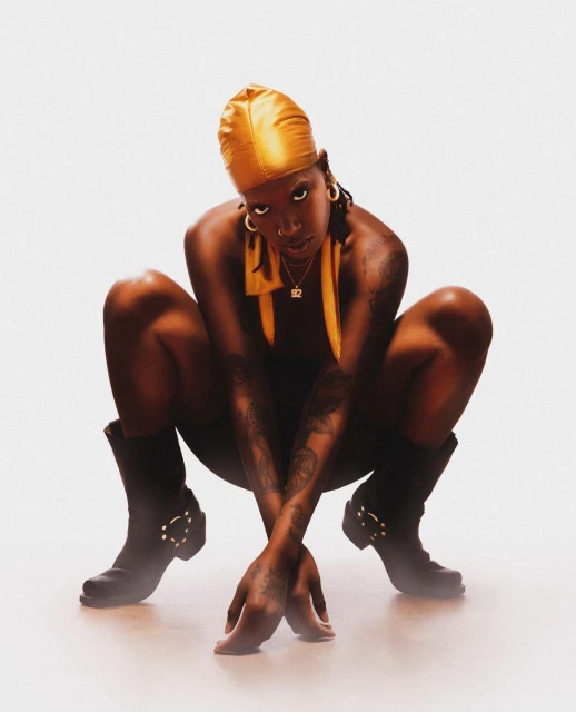 Woman with tattoos wearing a golden durag, necklace, and black boots crouches in a photo studio against a white backdrop.