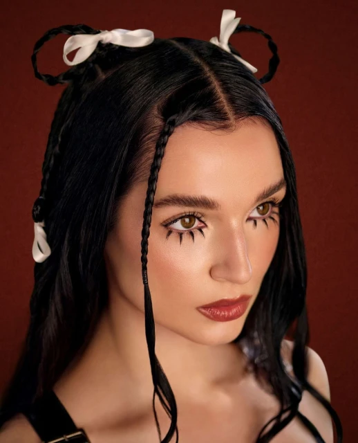 Close-up portrait of a fair-skinned woman with dark hair styled with braided pigtails and ribbons, plus dramatic eye makeup in a studio setting.
