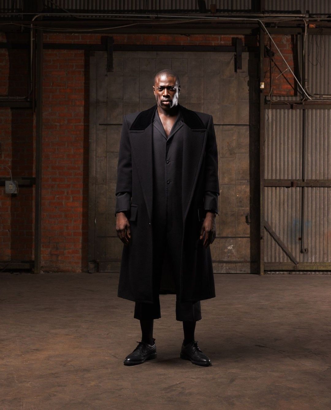 Fashionable Black man in dark coat posing in industrial space. He stands against a brick wall and metal door, wearing all black.