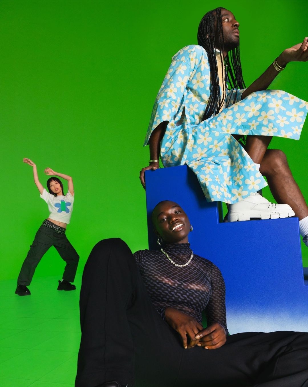 Three models pose in coordinated outfits against a bright green backdrop, featuring a blue block and bold fashion photography.