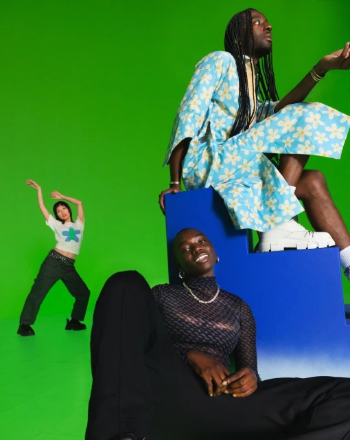 Three models pose in coordinated outfits against a bright green backdrop, featuring a blue block and bold fashion photography.