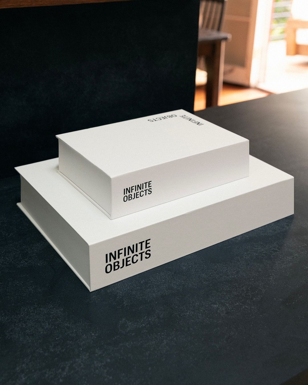 Two white rectangular boxes stacked on a black table with an out-of-focus view of a chair and partially open door in the background.