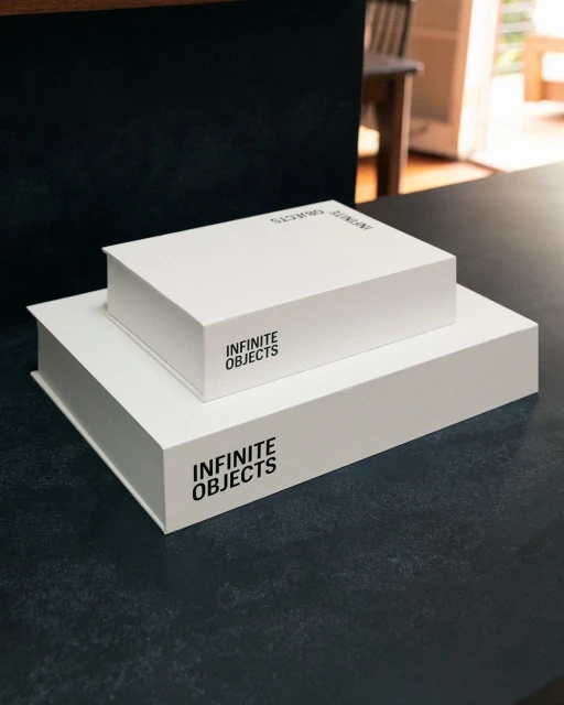 Two white rectangular boxes stacked on a black table with an out-of-focus view of a chair and partially open door in the background.