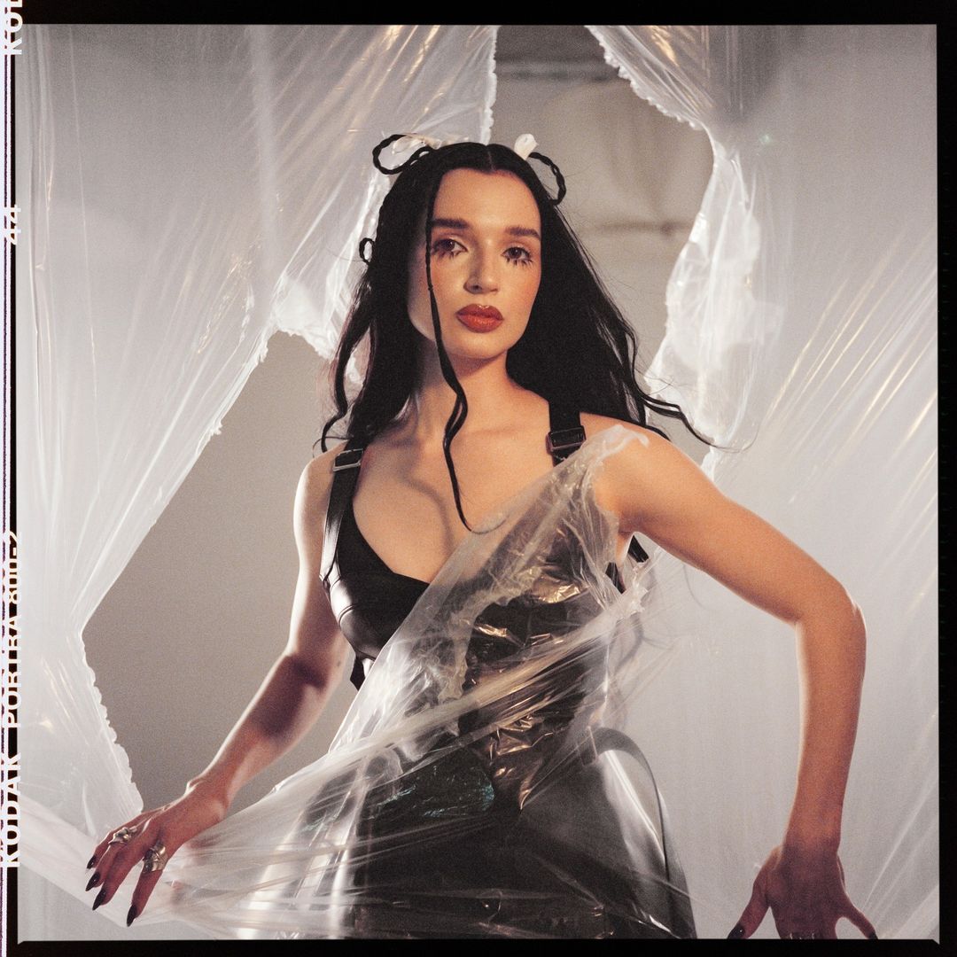 Adult female in a black dress and plastic wrap stands indoors, posing in front of a gray wall and abstract plastic sheeting backdrop.
