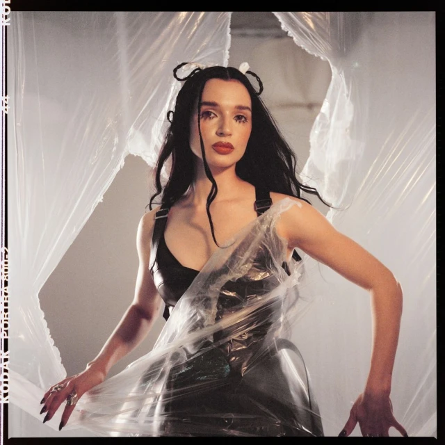 Adult female in a black dress and plastic wrap stands indoors, posing in front of a gray wall and abstract plastic sheeting backdrop.