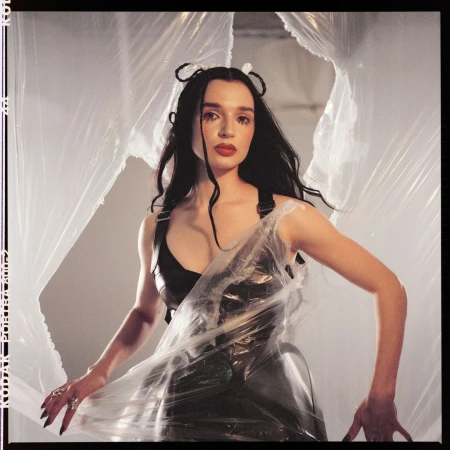 Adult female in a black dress and plastic wrap stands indoors, posing in front of a gray wall and abstract plastic sheeting backdrop.
