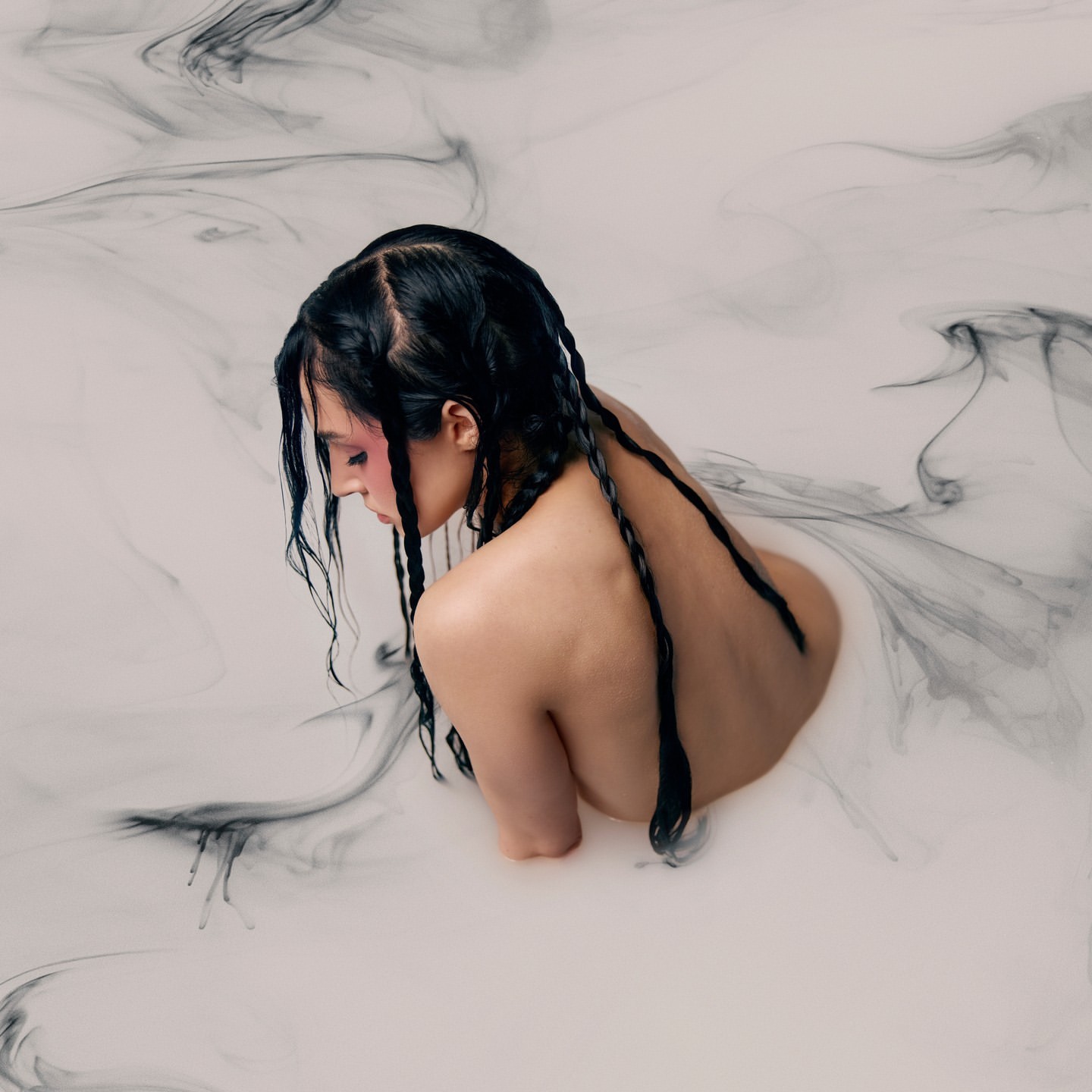 An adult female submerged in white liquid with black ink-like swirls, captured in a serene beauty shot featuring braided hair.