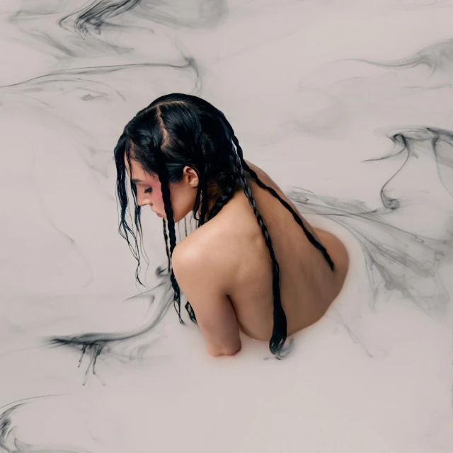 An adult female submerged in white liquid with black ink-like swirls, captured in a serene beauty shot featuring braided hair.