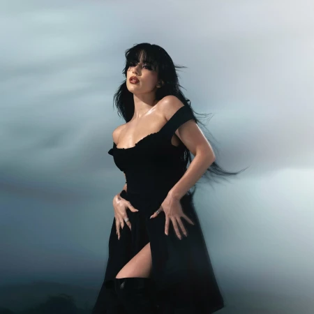 Woman with long dark hair wearing a black dress posing against an overcast sky