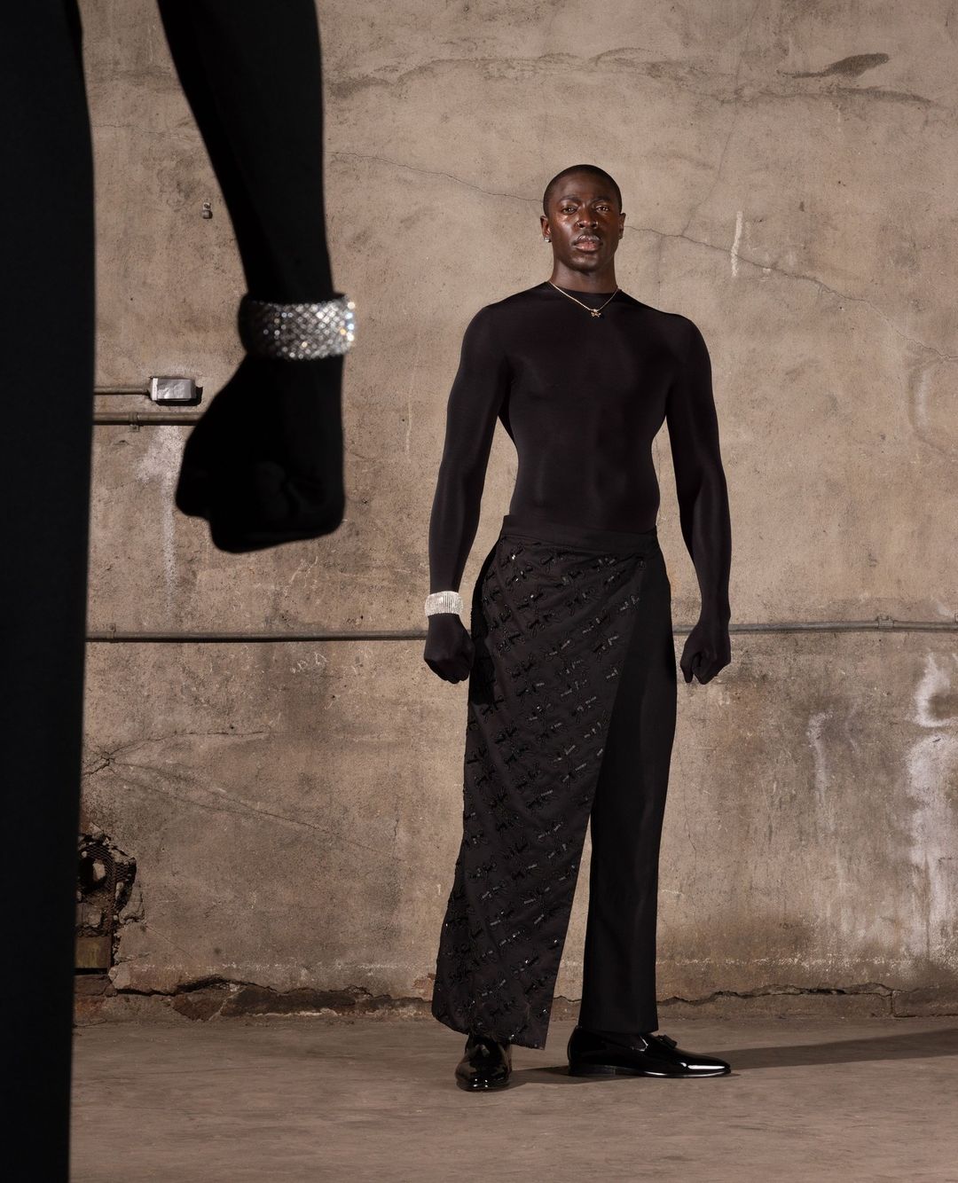 A well-lit fashion shot features a male model in black attire and jewelry, against a textured studio background with a partial figure.