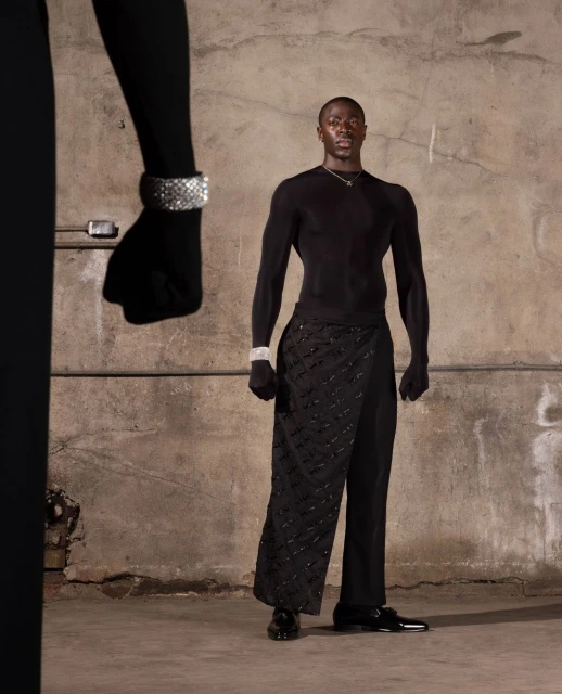 A well-lit fashion shot features a male model in black attire and jewelry, against a textured studio background with a partial figure.