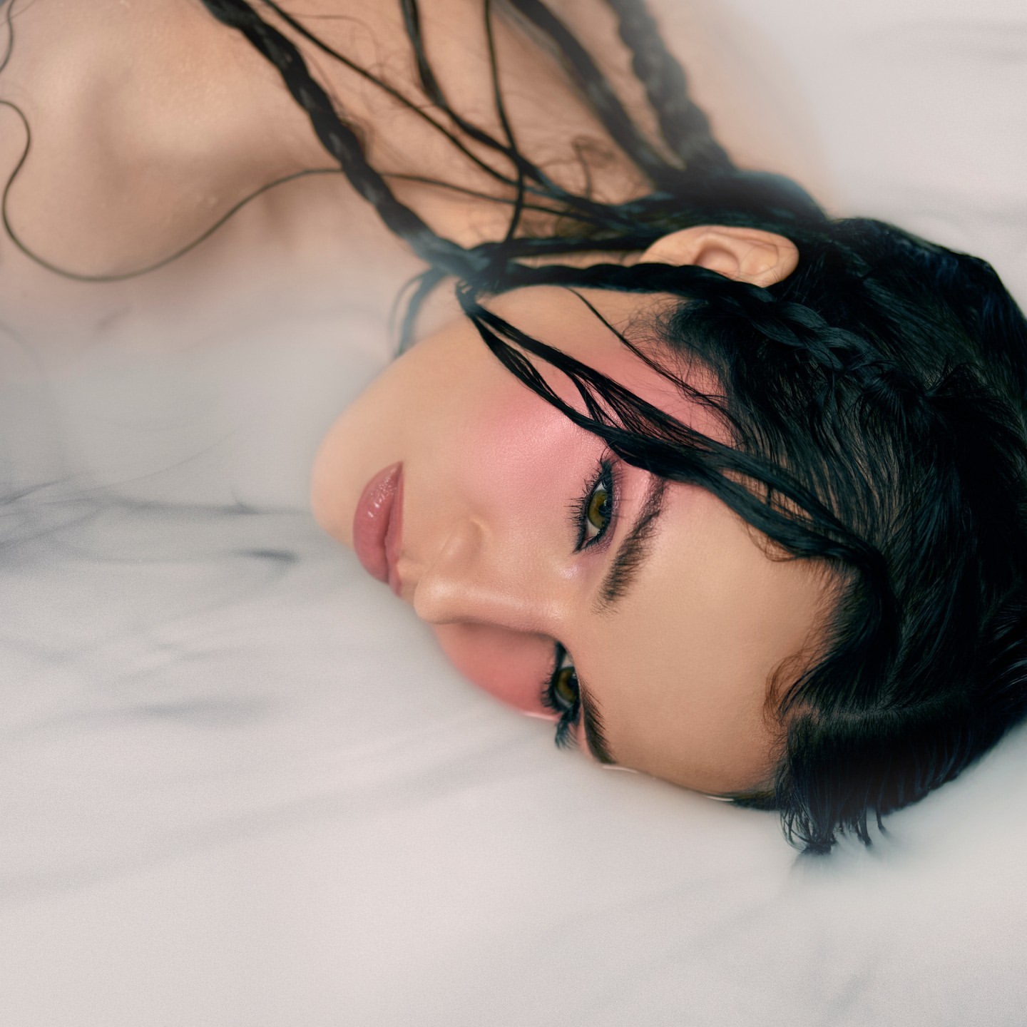 Close-up beauty shot of a woman with braided black hair, wet skin, and pink blush, lying back with a soft, dreamy aesthetic.