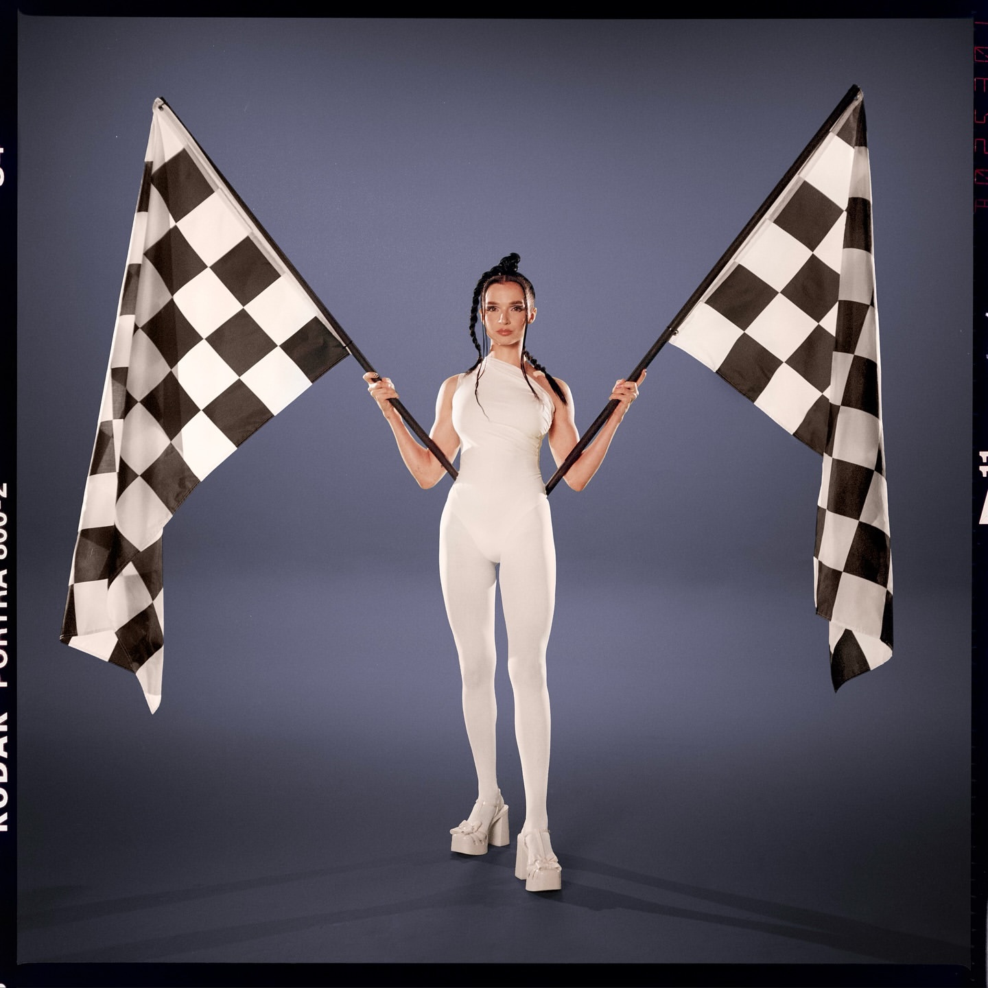 A woman in a white bodysuit stands in front of a gray background, holding two checkered racing flags, with a vintage film border.