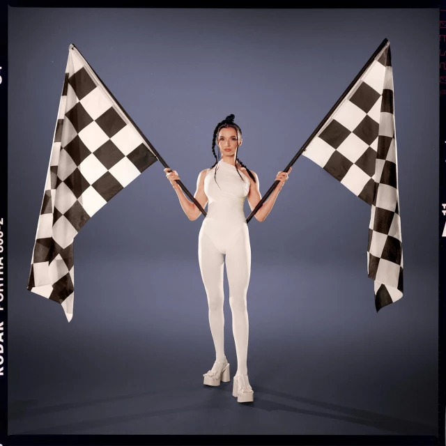 A woman in a white bodysuit stands in front of a gray background, holding two checkered racing flags, with a vintage film border.
