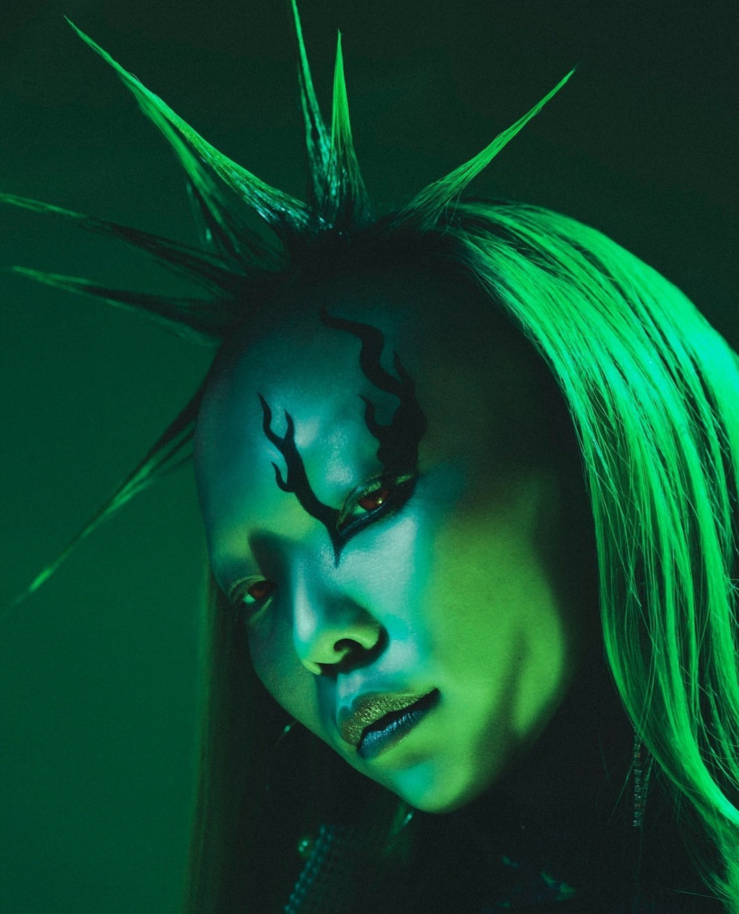 Close-up of an Asian woman with green hair and graphic makeup under green lighting in a beauty or editorial style portrait.