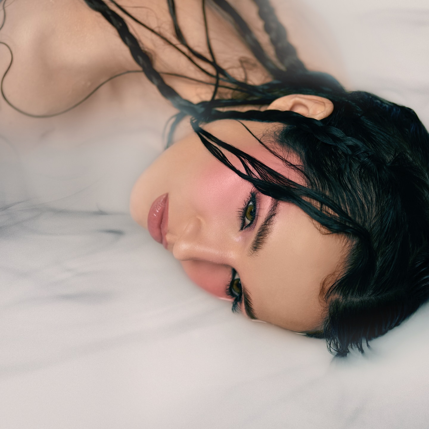 Glamorous beauty portrait: An adult woman with dark hair and pink blush lies in water, showcasing glossy lips and striking green eyes.