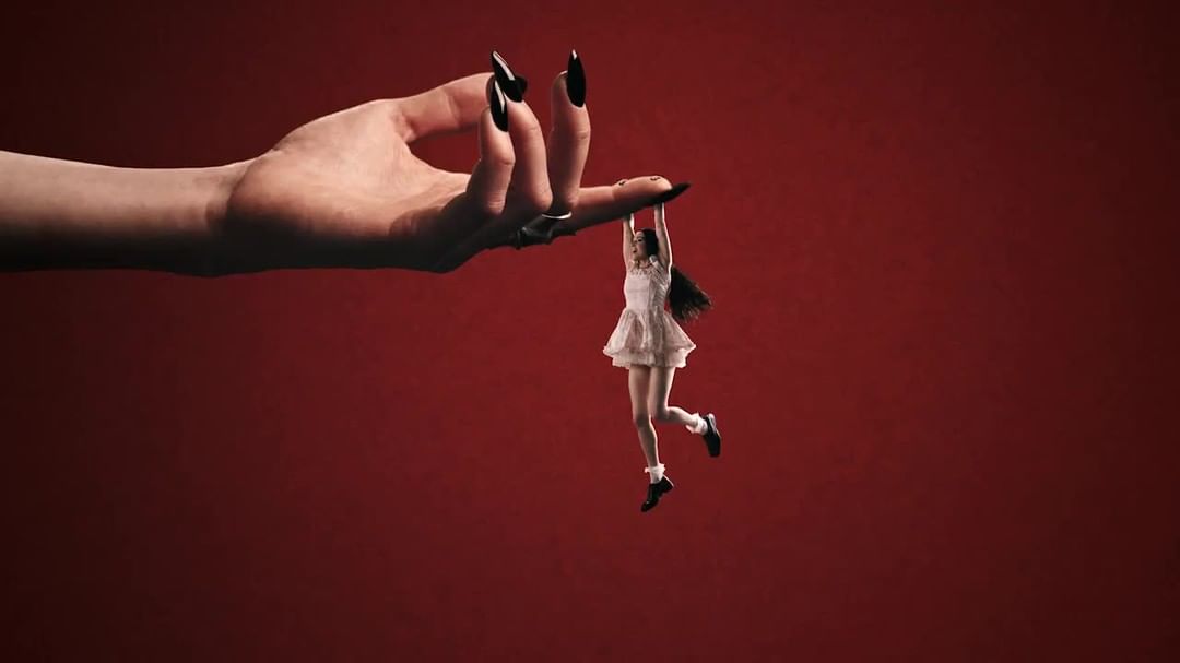 Conceptual image featuring a giant hand with long black nails holding a tiny woman in the air against a solid red background.