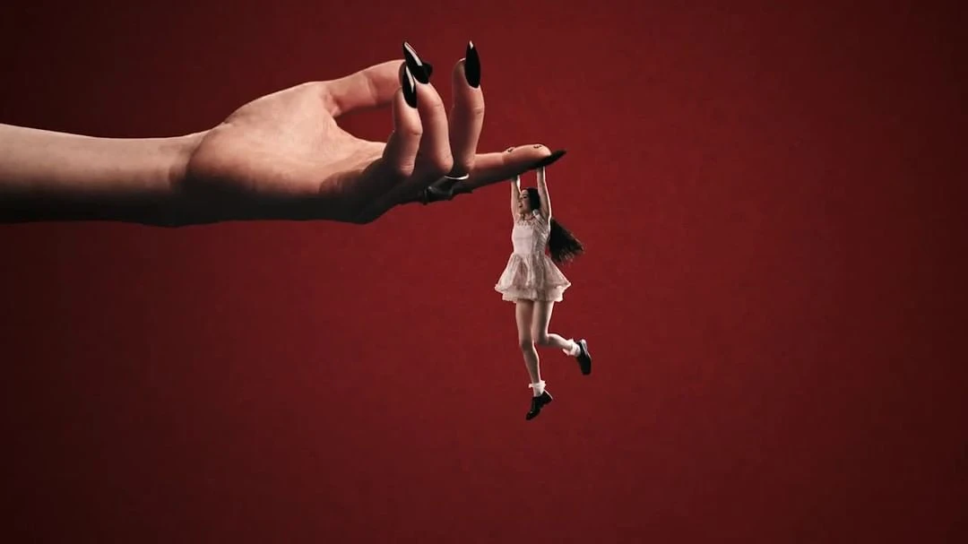 Conceptual image featuring a giant hand with long black nails holding a tiny woman in the air against a solid red background.