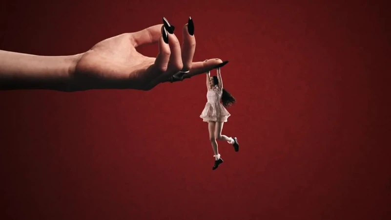 Conceptual image featuring a giant hand with long black nails holding a tiny woman in the air against a solid red background.