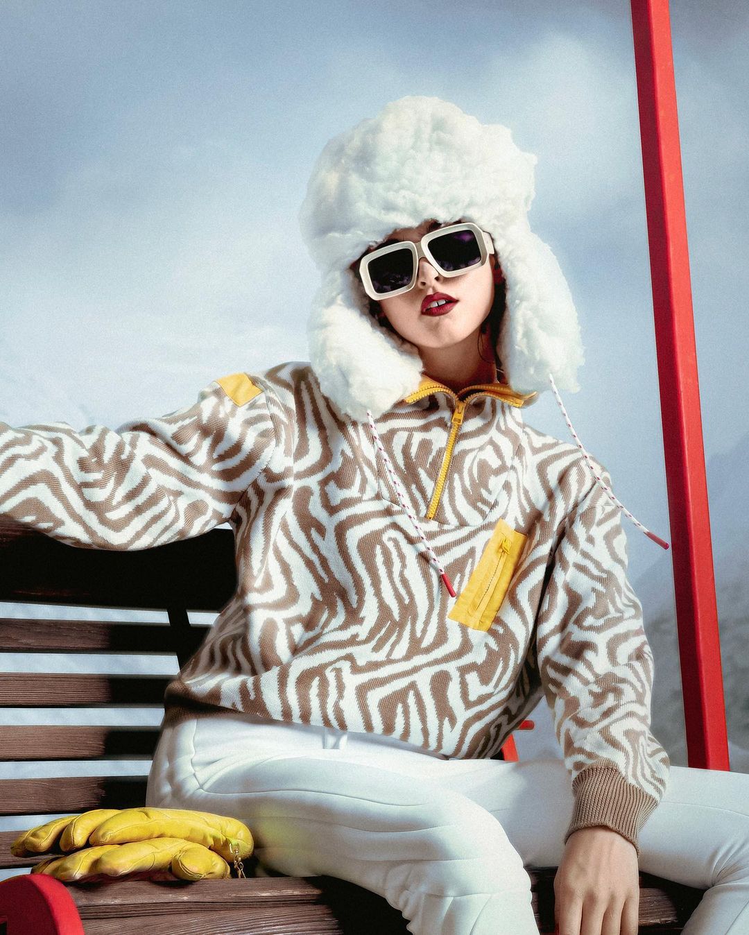 Fashion portrait of a woman in a zebra-print sweater, white pants, and a furry hat sitting on a bench against a cloudy sky backdrop.