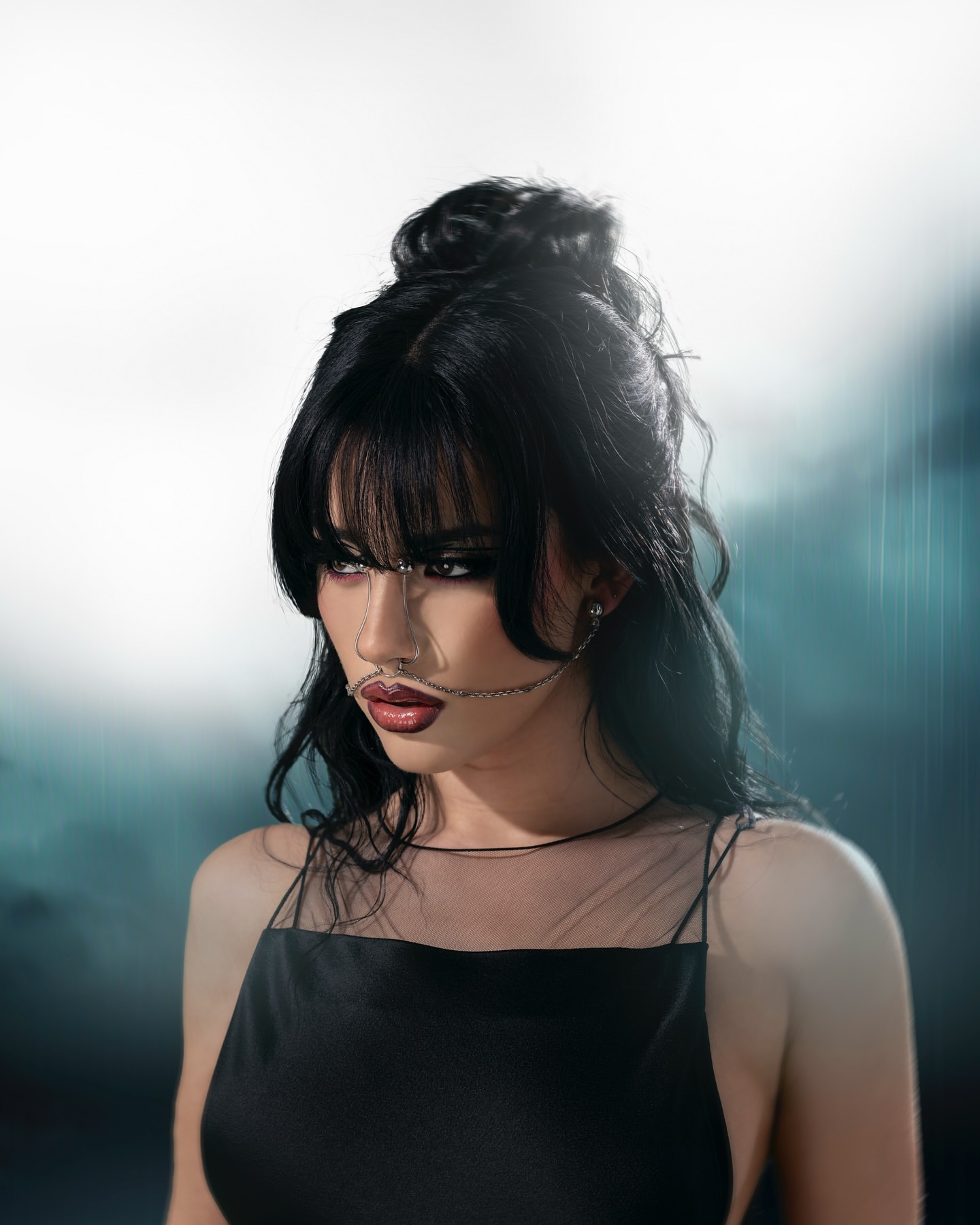 Portrait of a woman with dark hair in a topknot, wearing a black top and a face chain, posing against a dark, blurred background.