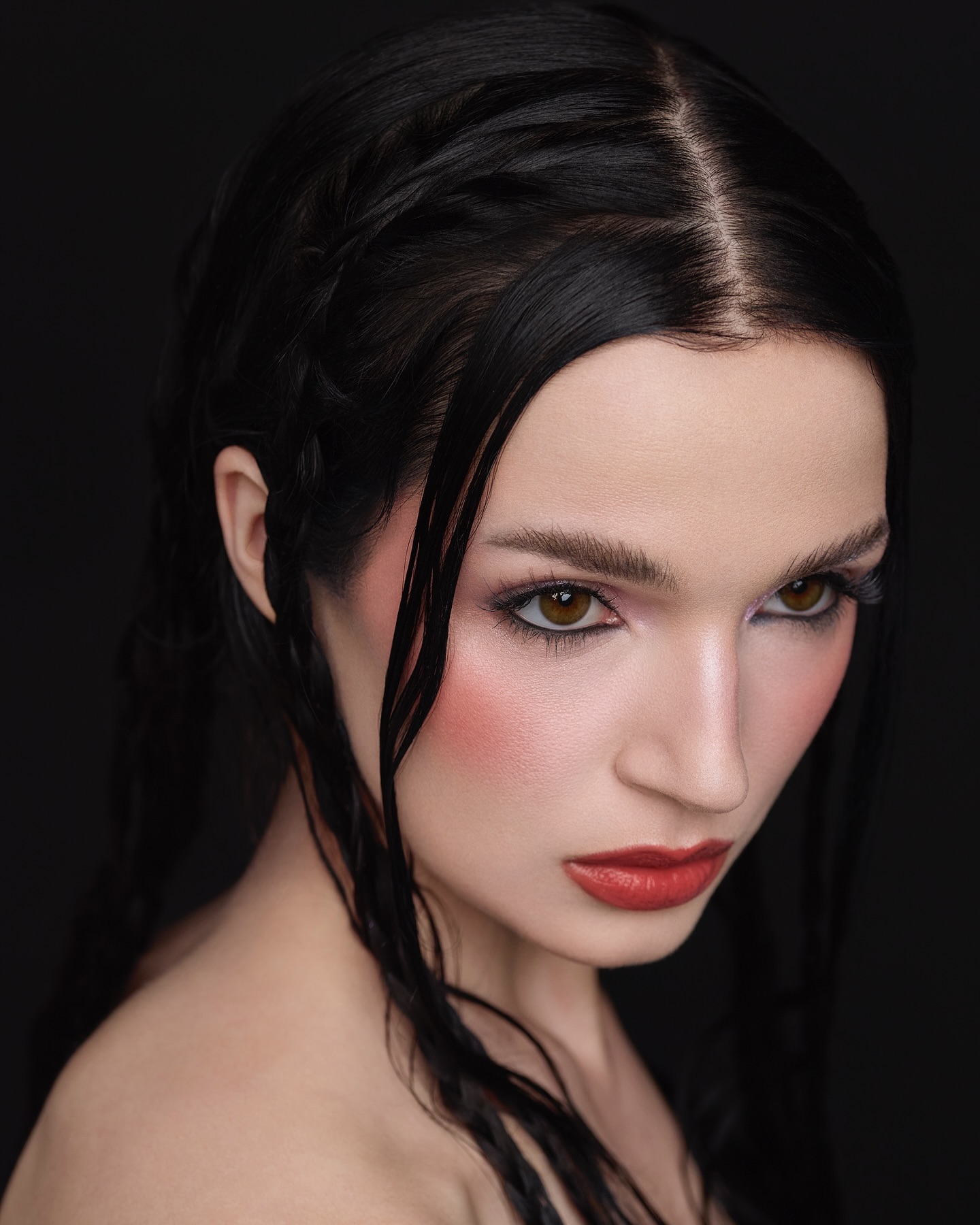 Headshot portrait of a woman with dark hair and bold makeup, styled with face blush and bright red lipstick against a black backdrop.
