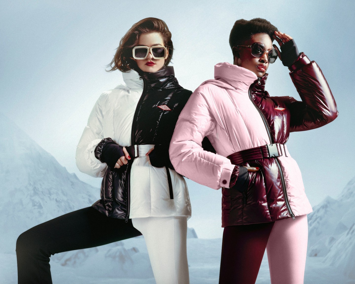 Two women pose outdoors on a snowy surface, wearing split-color ski jackets, sunglasses, and gloves in a fashion-style product shot.