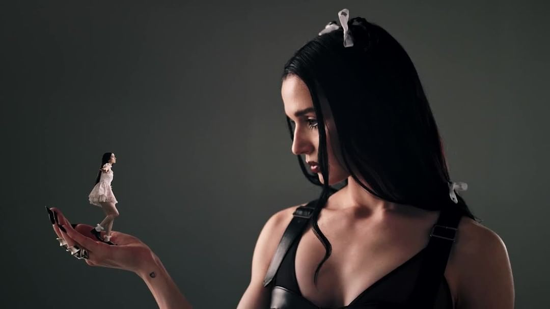 Woman with long black hair, dark eye makeup, and black top is holding a miniature woman in white dress on her hand.