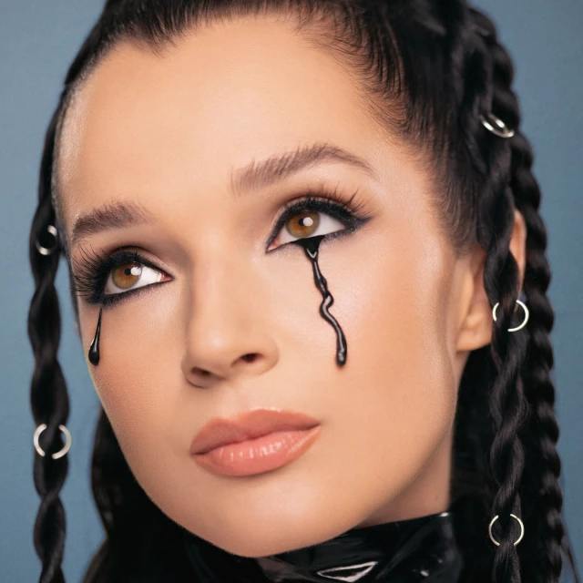 Close-up portrait of a young adult woman with dark hair braided with silver rings. She wears dark makeup and has black tears running down her face.