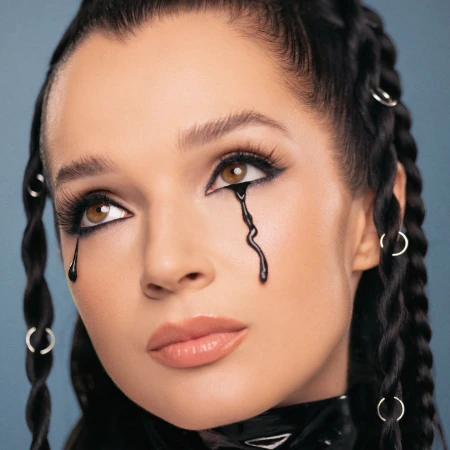 Close-up portrait of a young adult woman with dark hair braided with silver rings. She wears dark makeup and has black tears running down her face.