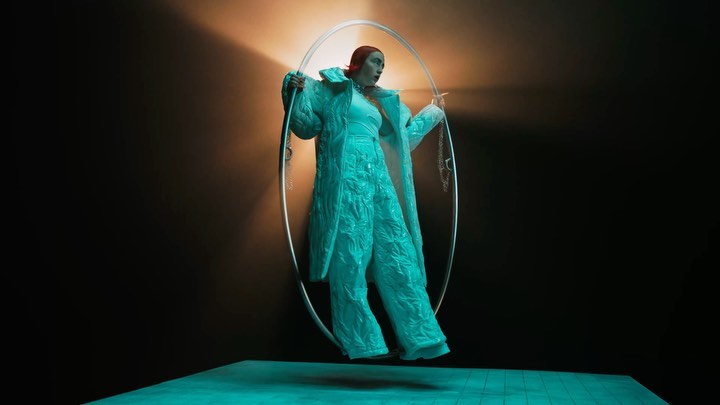 Woman in teal coat and trousers posing inside of a large circular frame against a dark background with orange and teal lighting.