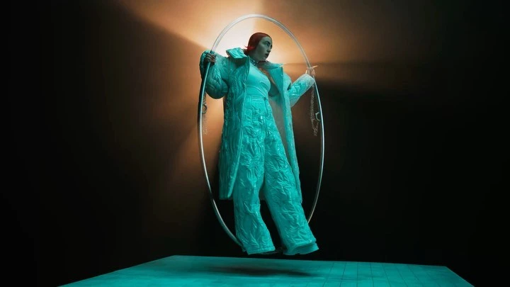 Woman in teal coat and trousers posing inside of a large circular frame against a dark background with orange and teal lighting.