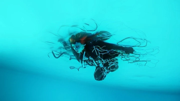 Woman with red highlights and black clothing surrounded by floating wires in a blue underwater environment in a conceptual fashion shot.