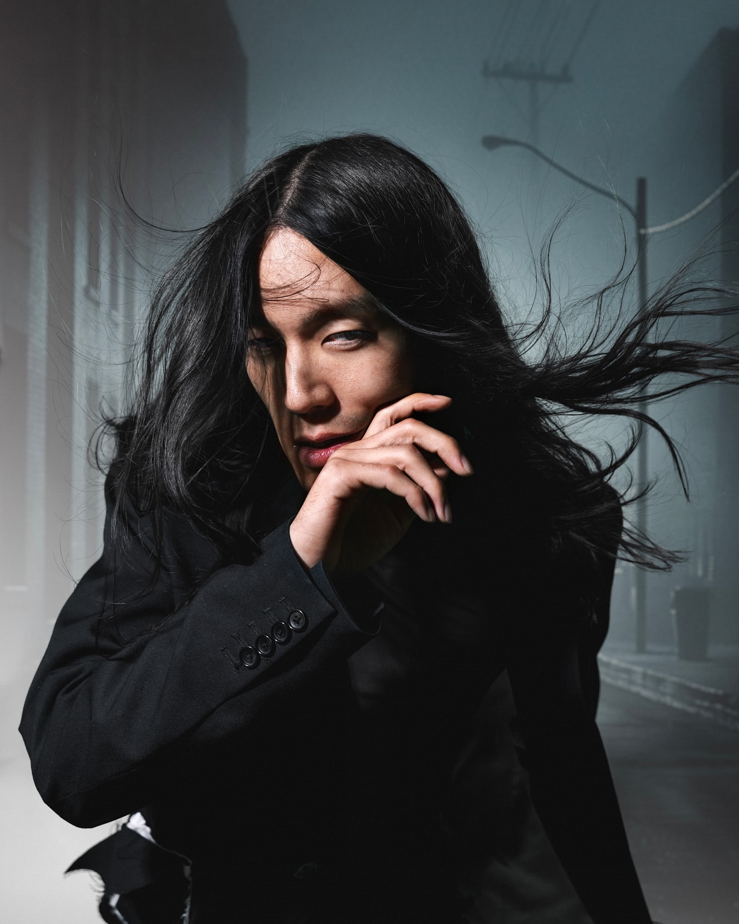 Portrait of an androgynous person with long black hair posing in a suit outdoors on a foggy city street at night, looking mysteriously.