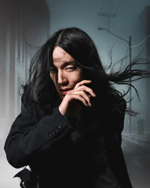 Portrait of an androgynous person with long black hair posing in a suit outdoors on a foggy city street at night, looking mysteriously.