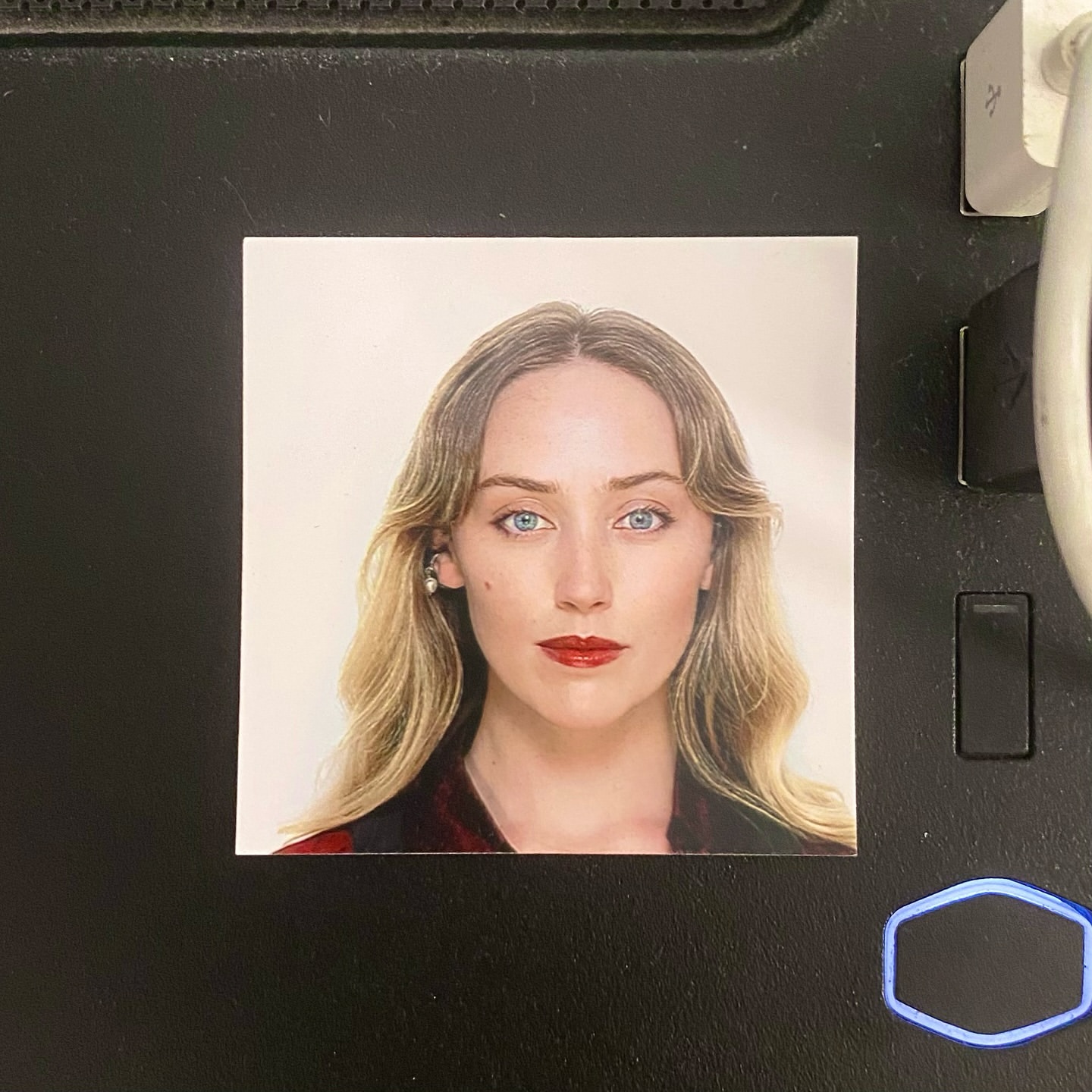 Headshot of a fair-skinned woman with long blonde wavy hair, blue eyes, and red lipstick, wearing a red and black patterned top.