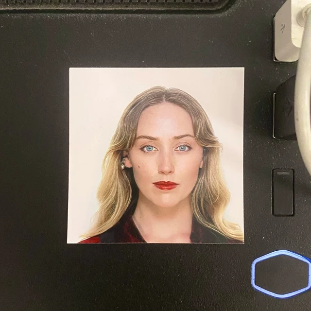 Headshot of a fair-skinned woman with long blonde wavy hair, blue eyes, and red lipstick, wearing a red and black patterned top.