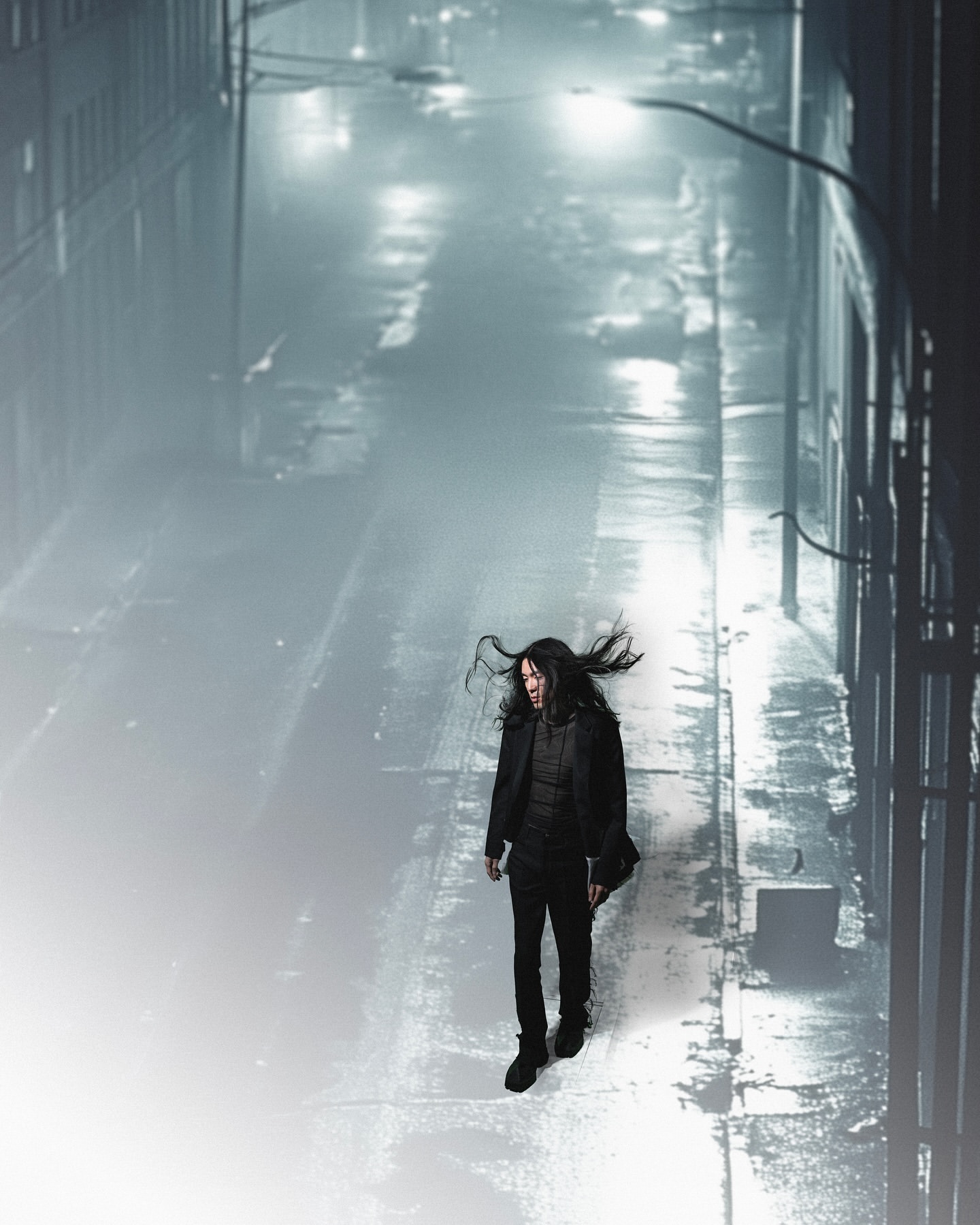 Adult male with long dark hair walks along wet urban alley at night, under streetlights, wearing dark jacket and pants in the fog.
