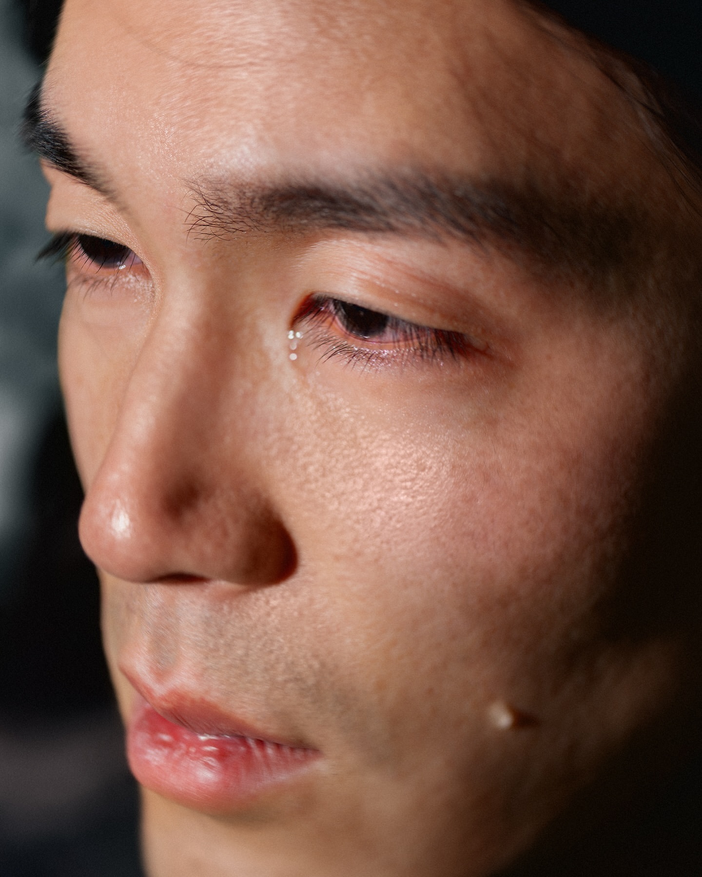 Close-up portrait of an Asian adult male with tears in his eyes, set against a dark, blurred background. Focus on skin texture and details.
